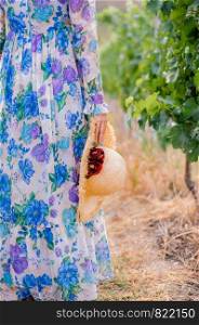 Woman in vaintage floral dress with natural coloured straw hat in hand walking along grape vine bush in vineyard - half body non recognize model