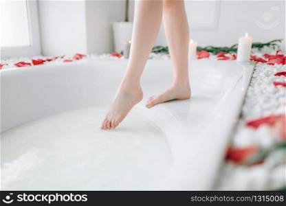 Woman in white bathrobe dips legs into the bath with foam. Luxury bathroom interior with window and palm branch decor. Woman in white bathrobe dips legs into the bath