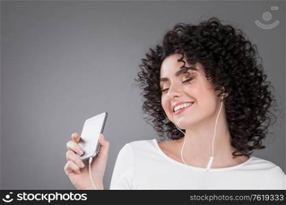 Woman in white dancing with earphones headphones listening to music on phone, gray background. Woman dancing with earphones