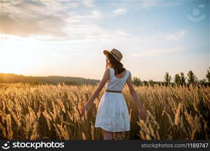 Woman in white dress and straw hat walking in wheat field on sunset. Female person on summer meadow, back view