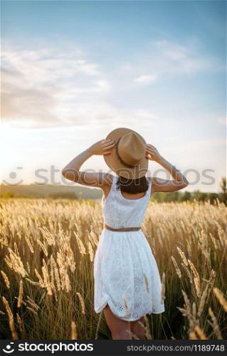 Woman in white dress and straw hat walking in wheat field on sunset. Female person on summer meadow. Woman in dress walking in wheat field on sunset