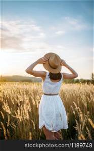 Woman in white dress and straw hat walking in wheat field on sunset. Female person on summer meadow. Woman in dress walking in wheat field on sunset