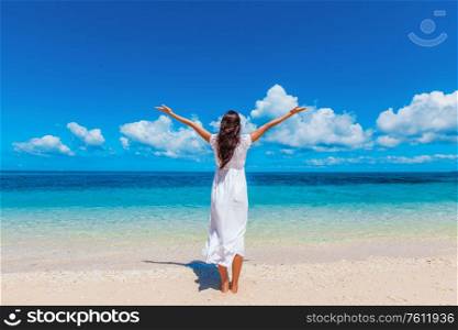 Woman in white dress posing in tropical sea beach with arms raised. Woman in dress walking on beach