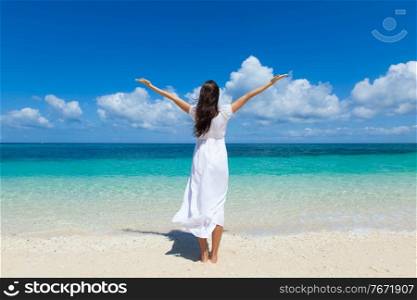 Woman in white dress posing in tropical sea beach with arms raised. Woman in dress walking on beach