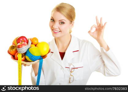 Woman in white lab coat holding fruits and colorful measure tapes isolated. Doctor dietitian recommending healthy food showing ok okay hand sign gesture. Diet.