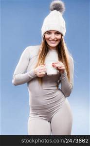 Woman in winter cap gray sports thermal underwear for skiing training holds mug with tea or coffee warming herself studio shot on blue. . girl in thermal underwear drinking tea