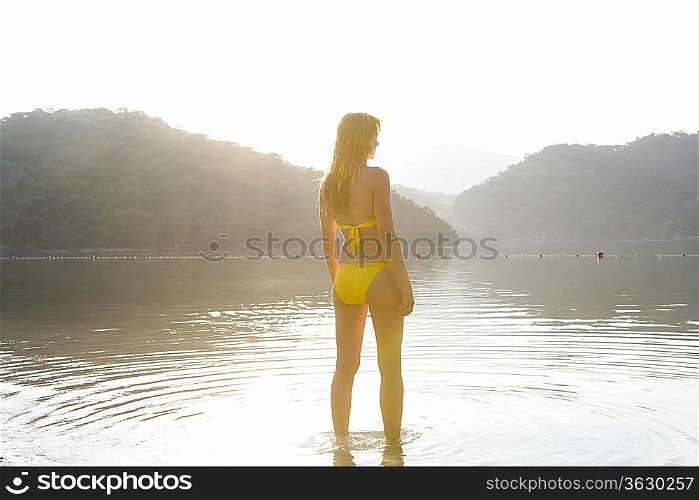 Woman in yellow bikini stands at edge of lake