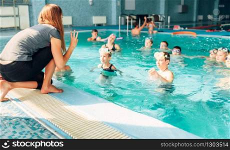 Woman instructor works with children in swimming pool. Kids with goggles in water listening female trainer. Happy kids in modern sport center. Concept of fun, leisure and recreation.. Instructor works with children in swimming pool