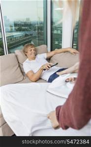 Woman ironing shirt while happy man watching TV on sofa at home