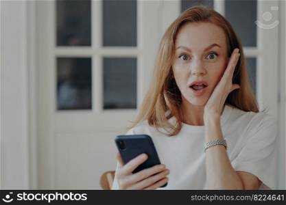 Woman is an excited customer holding phone. Surprised freelancer has deadline working from home. Redhead european lady gets shock. Concept of discount and profitable proposition and purchasing online.. Woman is an excited holding phone. Redhead european lady gets shock, freelancer has deadline.