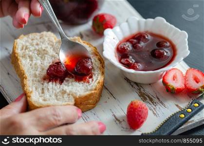 Woman is holding a spoon of strawberry jam and she is preparing breakfast in the kitchen.
