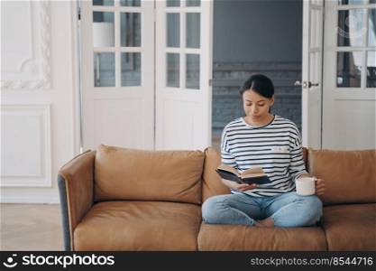 Woman is reading novel and drinks coffee or tea. Happy young european woman is reading book sitting on comfortable leather couch. Lady enjoying poetry. Living room interior with copy space.. Woman is reading novel and drinks coffee sitting on comfortable leather couch. Lady enjoying poetry.