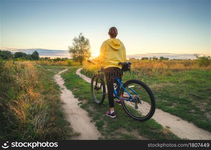 Woman is riding a mountain bike in cross country road at sunset in summer. Colorful landscape with sporty girl in yellow hoodie, bicycle, field, dirt road, green grass, trees, sky. Sport and travel