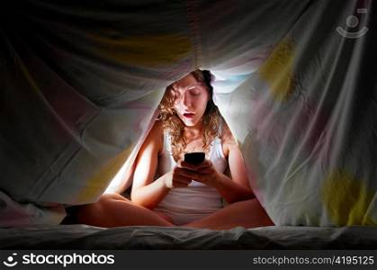 woman is sitting under cover in bed and chatting with friends via mobile phone