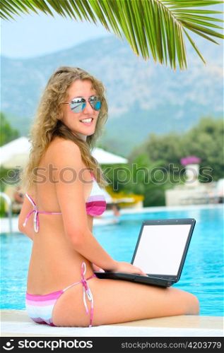woman is sitting with laptop on the edge of swimming pool under a palm tree