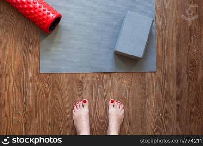 Woman is standing barefoot on floor in front of gymnastic mat and roller, she is going to do morning exercise complex. Only feet are visible. Healthy lifestyle, sport, weight loss concept. Horizontal.