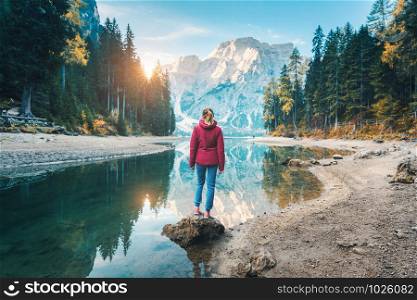 Woman is standing on the stone on Braies lake at sunrise in autumn. Dolomites, Italy. Landscape with girl, mountain, lake, beautiful reflection in water, trees, sky with sun. Forest in fall. Travel. Woman is standing on the coast of Braies lake at sunset