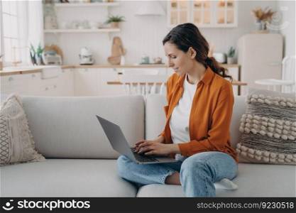 Woman is typing on pc and sitting on couch at home. European girl in orange shirt is chatting and browsing internet. Remote work or education. Businesswoman at cosy living room.. Woman is typing on pc and sitting on couch at home. European girl is chatting. Remote work.