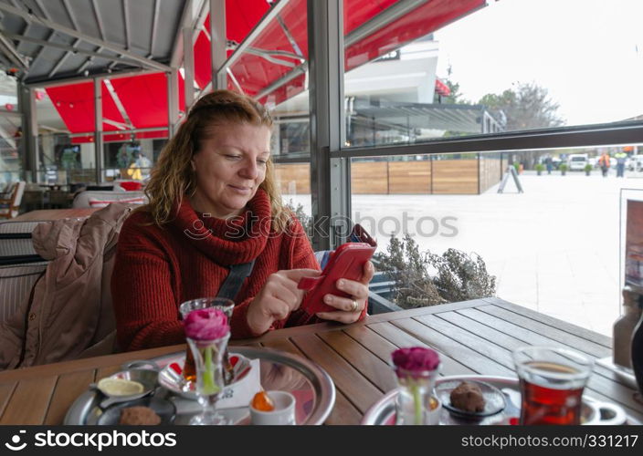 Woman is typing text message on smart phone in a cafe. Blond caucasian woman sitting at a table with a cup of tea using mobile phone.