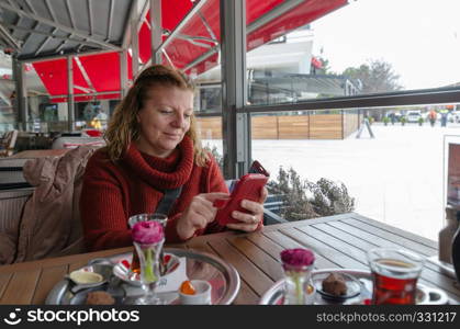 Woman is typing text message on smart phone in a cafe. Blond caucasian woman sitting at a table with a cup of tea using mobile phone.