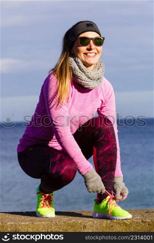 Woman jogger wearing warm sporty clothes tying running shoes while jogging outdoor on seaside by cold day. Sport healthy lifestyle. Woman jogger tying running shoes outdoor