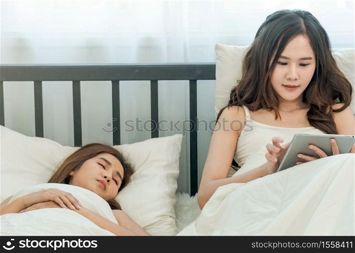 Woman just woke up and laying on the bed using tablet or video call while her friend still sleeping. Tablet , Gadget, Technology, Shopping, Friend, Friendship, LGBT, lesbian, gay, bisexual.
