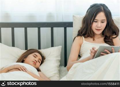Woman just woke up and laying on the bed using tablet or video call while her friend still sleeping. Tablet , Gadget, Technology, Shopping, Friend, Friendship, LGBT, lesbian, gay, bisexual.