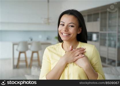 Woman keeps hands close to heart. Conceptual portrait of young happy spanish woman showing love. Advertising banner mockup. Handsome girl in yellow shirt in living room at home. Modern interior.. Woman keeps hands close to heart. Conceptual portrait of young happy woman showing love.