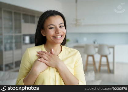 Woman keeps hands close to heart. Conceptual portrait of young happy spanish woman showing love. Advertising banner mockup. Handsome girl in yellow shirt in living room at home. Modern interior.. Woman keeps hands close to heart. Conceptual portrait of young happy woman showing love.