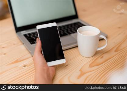woman laptop computer smartphone mug office supplies technological devices. Young lady holding a smartphone with left hand. Gray laptop computer on the background with black keyboard and a mug of coffee. Office supplies, technological devices and wooden desk.