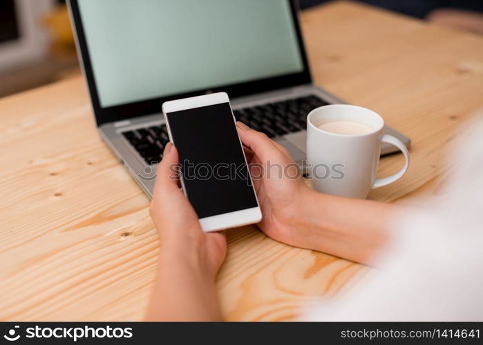 woman laptop computer smartphone mug office supplies technological devices. Young lady holding a smartphone with both hands. Gray laptop computer on the background with black keyboard and a mug of coffee. Office supplies, technological devices and wooden desk.