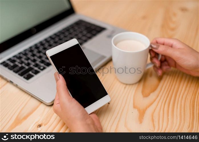 woman laptop computer smartphone mug office supplies technological devices. Young lady holding a smartphone with left hand. Gray laptop computer on the background with black keyboard, a mug of coffee, office supplies, technological devices and wooden desk.