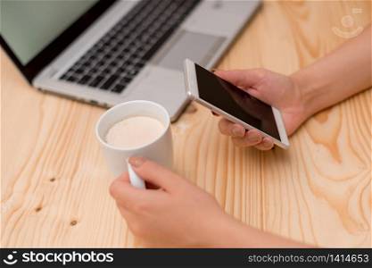 woman laptop computer smartphone mug office supplies technological devices. Young lady holding a smartphone and mug of tea. Gray laptop computer on the background with black keyboard a cup of coffee. Office supplies, technological devices and wooden desk.