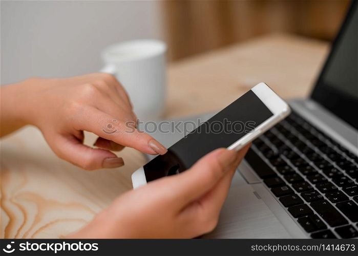 woman laptop computer smartphone mug office supplies technological devices. Young lady using a smartphone and touching the screen. Gray laptop computer on the background with black keyboard and a mug of coffee. Office supplies, technological devices and wooden desk.