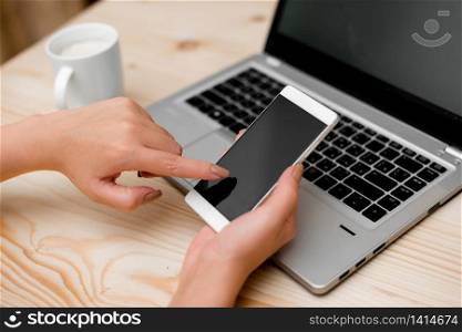 woman laptop computer smartphone mug office supplies technological devices. Young lady using a smartphone and touching the screen. Gray laptop computer on the background with black keyboard and a mug of coffee. Office supplies, technological devices and wooden desk.