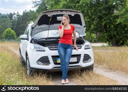 Woman leaning on broken car in field and calling for help on mobile phone