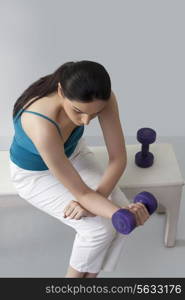 Woman lifting dumbbells while sitting on bench against gray background