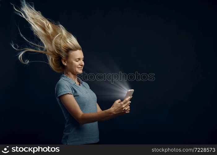 Woman looking on phone against powerful airflow in studio, back view, developing hairstyle effect. Female person and wind, lady isolated on dark background. Woman looking on phone against powerful airflow