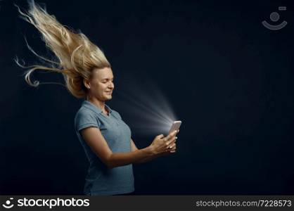 Woman looking on phone against powerful airflow in studio, back view, developing hairstyle effect. Female person and wind, lady isolated on dark background. Woman looking on phone against powerful airflow
