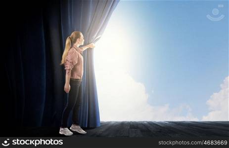 Woman looking out from curtain. Young woman in casual opening blue curtain and looking at clear sky
