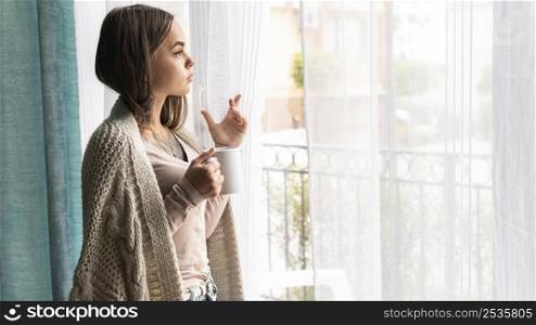 woman looking through window home during pandemic while having coffee