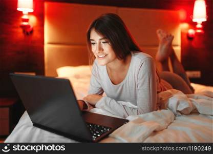 Woman lying in bed and looking on laptop screen, good morning, bedroom interoir on background. Lady awake. Woman lying in bed and looking on laptop screen