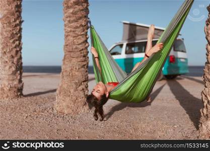 Woman lying in hammock between palm trees on the beach with a camper van in the background. Woman lying in hammock between palm trees on the beach