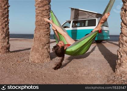 Woman lying in hammock between palm trees on the beach with a camper van in the background. Woman lying in hammock between palm trees on the beach