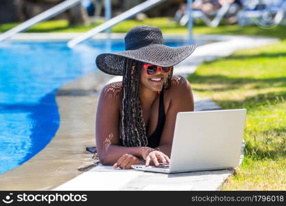 Woman lying on the edge of the pool with a laptop . African American woman with hat and sunglasses enjoying a summer day.. Woman lying on the edge of the pool with a laptop