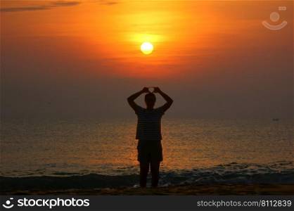 woman making heart pattern by hand while standing on beach looking sun set