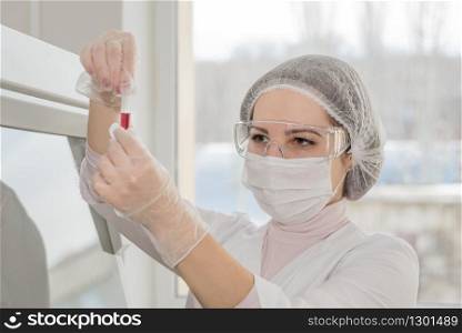 Woman medic in a white protective clothing conducts research with blood in a test tube in a real lab