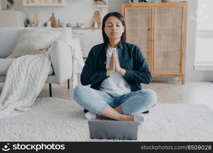 Woman meditate at laptop while online yoga lesson, sitting in lotus asana on floor at home. Calm female listen audio affirmations feels serenity. Wellness, calmness, stress relief concept.. Woman practice yoga, meditate at laptop in lotus asana on floor at home. Wellness, stress relief