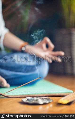 Woman meditating in lotus position on turquoise yoga mat on wooden floor. Focus on incense stick and smoke. Unrecognizable yoga practitioner in the background. Relax after Yoga training