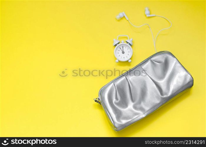 Woman modern morning concept. Silver cosmetic bag, alarm clock and headphones on yellow background. Colors of the year 2021 - ultimate gray and illuminating.. Woman modern morning concept. Silver cosmetic bag, alarm clock and headphones on yellow background. Colors of year 2021 - ultimate gray and illuminating.